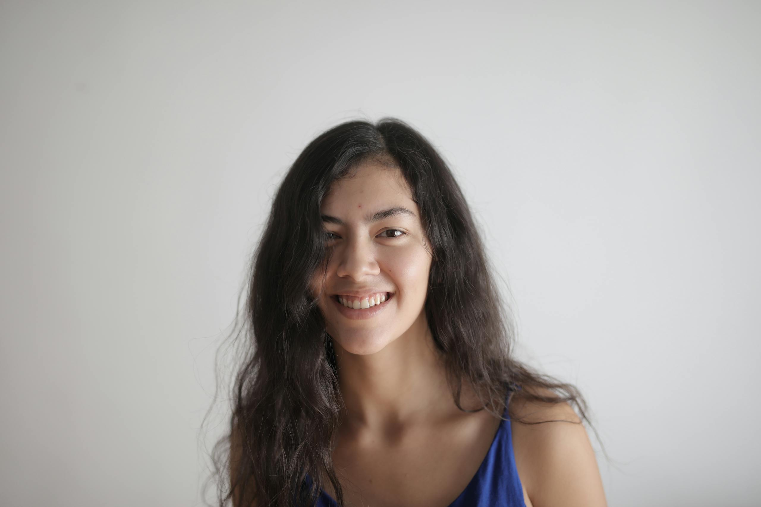 A cheerful woman with long hair smiles warmly in a blue top against a white background.
